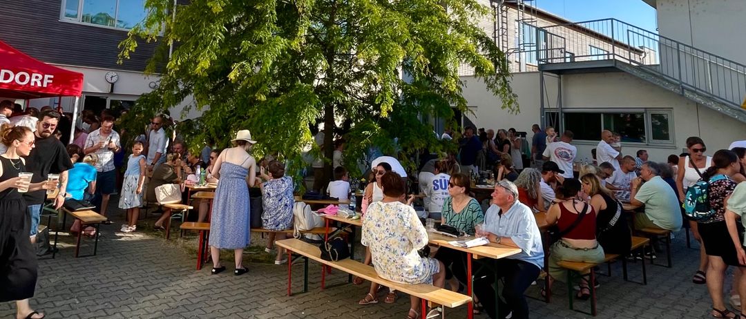 Eine Gruppe von Menschen sitzt unter einem Baum um Tische herum, mit Gebäuden im Hintergrund und einem roten Zelt auf der linken Seite. Sie tragen Freizeitkleidung und einige tragen Hüte.