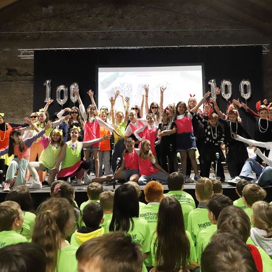 Eine Gruppe von Kindern in neongrünen Shirts tritt auf der Bühne auf und hebt die Hände. Dahinter tanzen Menschen in bunten Kostümen. Eine Projektionsleinwand zeigt die Zahl 100.
