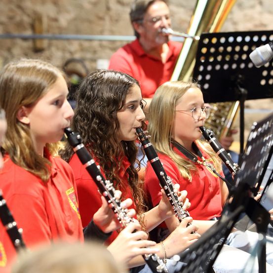 Drei Mädchen in roten Shirts spielen Klarinetten während einer Aufführung, mit einem Mann, der hinter ihnen Trompete spielt.
