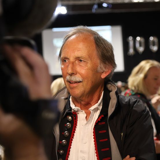 Ein älterer Mann mit Schnurrbart lächelt und schaut in die Kamera. Er trägt eine schwarz-weiße Jacke. Hinter ihm ist ein verschwommenes Bild einer Frau mit blonden Haaren. Es gibt einen schwarzen Hintergrund mit der Nummer 100.