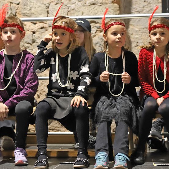 Fünf Kinder sitzen auf einer Holzbank und tragen rote Stirnbänder und Perlenketten. Sie scheinen an einer Schulveranstaltung teilzunehmen. Hinter ihnen ist eine Steinmauer und ein Metallgeländer zu sehen.