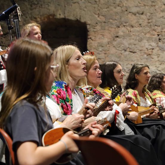 Eine Gruppe von Frauen in farbenfrohen traditionellen Kleidungsstücken spielt Saiteninstrumente. Sie sitzen auf Stühlen, einige singen, während andere sich auf ihre Instrumente konzentrieren. Hinter ihnen spielt ein Mann Cello.