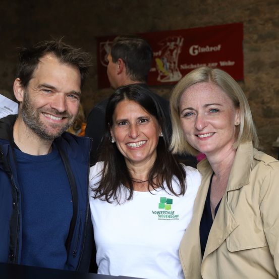 Drei Menschen stehen lächelnd für ein Foto zusammen. Ein Mann mit Bart, eine Frau in einem weißen T-Shirt und eine weitere Frau in einer beigen Jacke. Hinter ihnen ist ein Banner mit dem Wort 'Gudruf'.