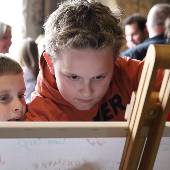 Zwei Jungen konzentrieren sich auf eine Tafel mit Schriftzügen, vor einem hölzernen Staffelei. Hinter ihnen schauen Leute in Freizeitkleidung zu. Der Junge auf der linken Seite trägt ein weißes T-Shirt.