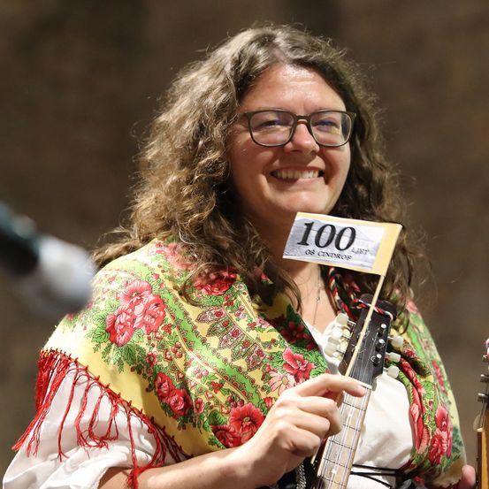 Eine Frau mit lockigem Haar lächelt und hält eine Gitarre. Sie trägt ein farbenfrohes Blumenkleid und eine Brille. Sie hält eine Karte mit der Nummer 100.