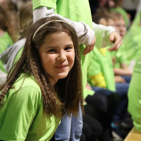 Ein junges Mädchen mit Stirnband lächelt in die Kamera, umgeben von anderen in grünen Shirts.