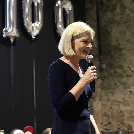 Eine Frau in einem blauen Kleid lächelt und hält ein Mikrofon, vermutlich hält sie eine Rede. Hinter ihr befinden sich drei große Folienballons mit der Nummer 10. Einige Menschen sind im Publikum, und eine Ziegelmauer ist im Hintergrund.