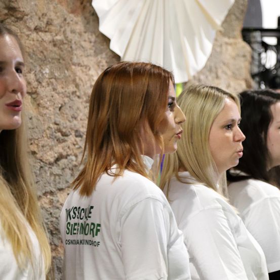 Eine Gruppe von Frauen in weißen T-Shirts mit dem Text 'YKSSCUE SIEÐORF OSNOVAKINDROF' steht zusammen. Sie stehen vor einer Steinmauer mit einem großen weißen Fächer im Hintergrund.