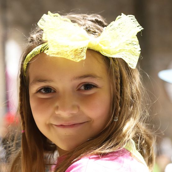 Ein Nahaufnahme eines jungen Mädchens mit braunen Haaren, das ein rosa Shirt und einen gelben Bogen im Haar trägt. Sie lächelt, und der Hintergrund ist unscharf.