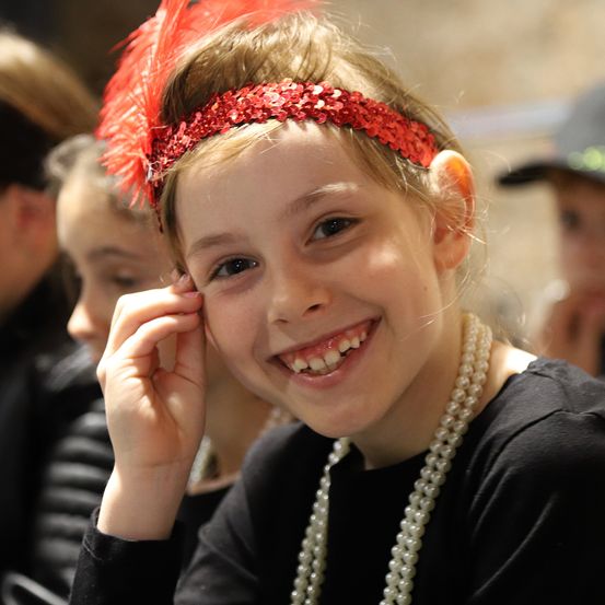 Ein junges Mädchen in einem schwarzen Shirt mit einer roten Pailletten-Stirnband, lächelt und berührt ihre Wange.