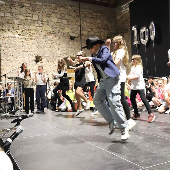 Eine Gruppe von Menschen tanzt auf der Bühne. Ein Mann in Anzug und Hut führt den Tanz an. Es sind Kinder mit Sonnenbrillen. Dahinter ist eine Steinmauer mit Luftballons.