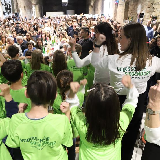 Eine Gruppe von Menschen in grünen Shirts tanzt in einer Halle. Im Hintergrund sind viele Menschen versammelt, einige machen Fotos. Zwei Frauen in weißen Shirts führen den Tanz an.