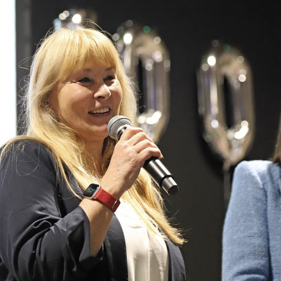 Eine Frau mit blonden Haaren spricht in ein Mikrofon. Sie trägt eine schwarze Jacke und ein weißes Hemd. Hinter ihr befinden sich zwei große silberne Nullen.