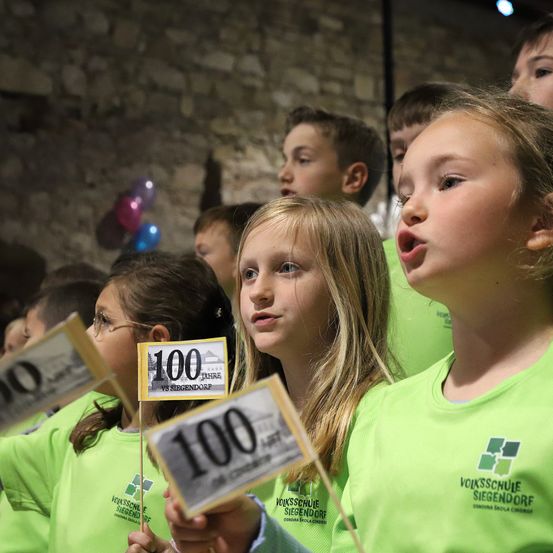Kinder in grünen T-Shirts, die Schilder mit der Aufschrift '100' halten, singen zusammen. Dahinter steht eine Gruppe von Kindern in einem Raum mit einer Ziegelsteinwand und Luftballons.
