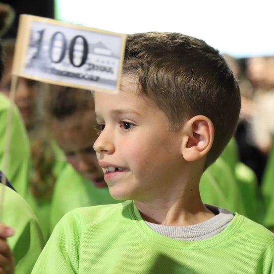 Ein junger Junge in einem grünen Shirt hält ein kleines Schild mit der Nummer 100. Hinter ihm sind andere Kinder in grünen Shirts zu sehen.