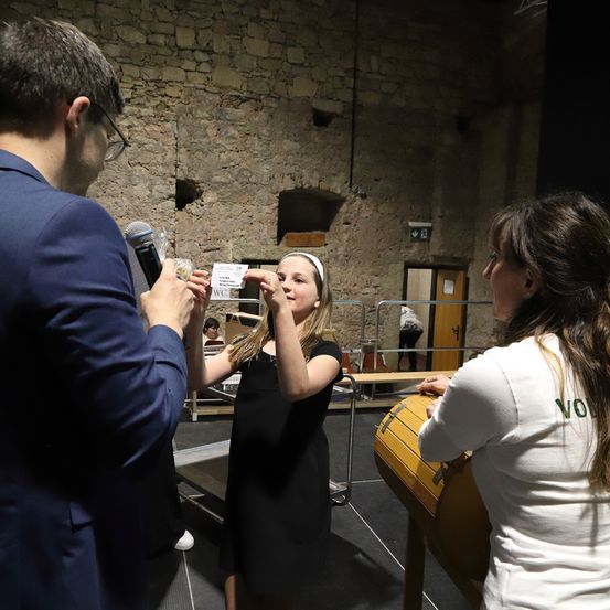 Ein Mann in einem Anzug spricht in ein Mikrofon, während eine Frau ein Stück Papier hält und aus einem Becher trinkt. Eine andere Frau schaut sie an. Sie befinden sich in einem Raum mit einer Backsteinmauer und einem Trommel.