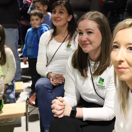 Drei Frauen in weißen Hemden sitzen in einem Raum mit einer Menge. Eine Frau trägt eine Uhr und hat ihre Hände gefaltet. Hinter ihnen sind Kinder und Erwachsene anwesend, einige stehen.