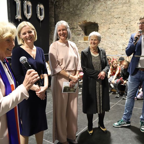 Fünf Erwachsene stehen auf einer Bühne mit einer Menschenmenge im Hintergrund. Eine Frau hält ein Mikrofon. Dahinter ist eine große Nummer 100 zu sehen.