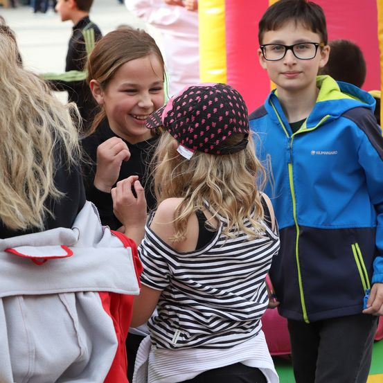 Drei Kinder stehen in der Nähe eines aufblasbaren Spielbereichs und lächeln. Ein Mädchen in einem schwarz-weiß gestreiften Shirt und ein Junge in einem blauen Mantel interagieren miteinander.