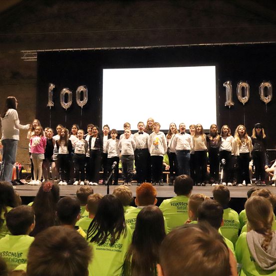 Ein Chor von Kindern in passenden grünen Shirts tritt auf der Bühne vor einem Publikum auf. Dahinter schweben zwei silberne Luftballons mit der Nummer 100.