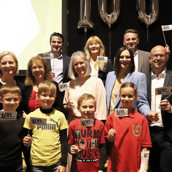 Eine Gruppe von Menschen steht auf einer Bühne mit Luftballons und Schildern mit der Aufschrift 100. Sie alle tragen Lächeln. Es gibt Kinder, Erwachsene und einen Erwachsenen mit einem Gips auf seinem Arm.