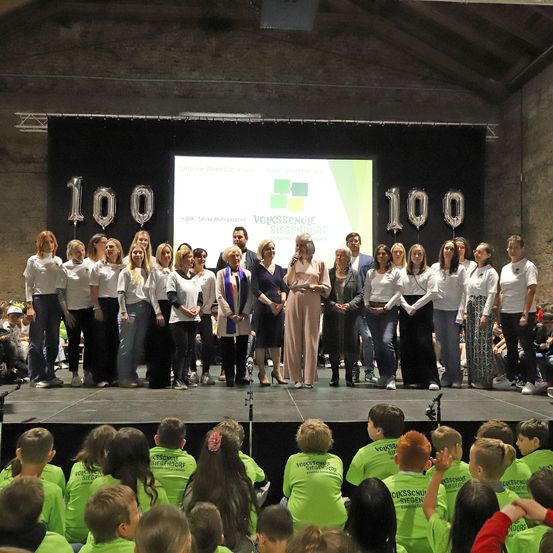 Eine Gruppe von Menschen, einschließlich Kinder in grünen Shirts, steht auf einer Bühne mit Luftballons und einer Leinwand. Sie feiern wahrscheinlich einen Meilenstein.