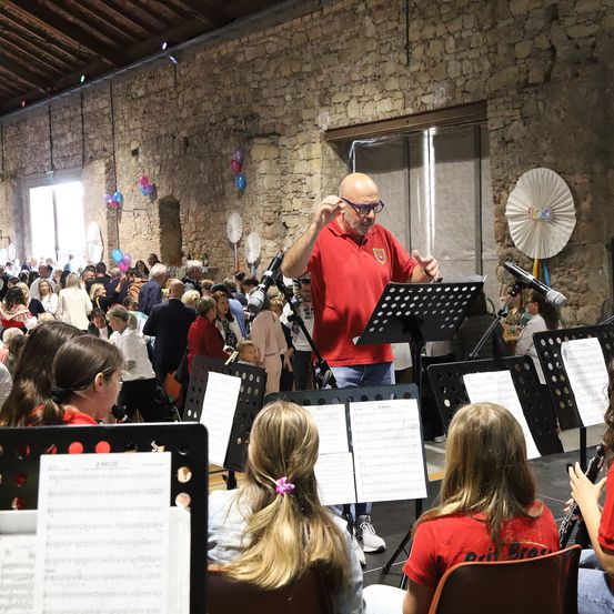 Ein älterer Mann in einem roten Hemd steht vor einem Mikrofon und leitet eine Band. Hinter ihm sitzen Kinder und spielen Instrumente. Im Hintergrund steht oder sitzt eine Menschenmenge und beobachtet die Aufführung. Luftballons und Ventilatoren hängen an der Steinmauer.