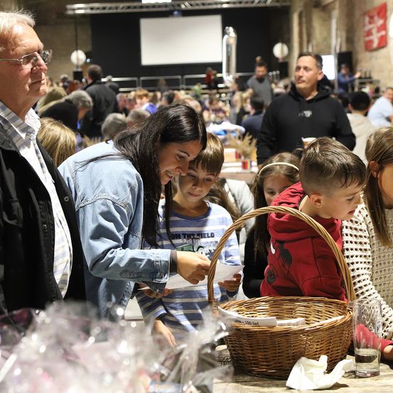 Mehrere Menschen versammeln sich in einem Raum, wahrscheinlich für eine Veranstaltung. Eine Frau und ein Junge betrachten Gegenstände in einem Korb auf einem Tisch.