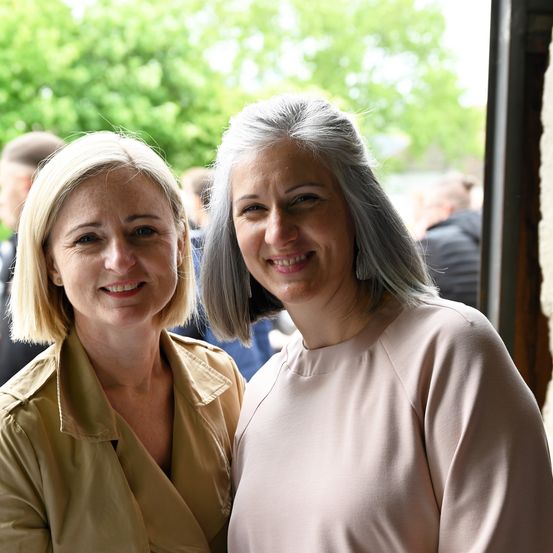 Zwei Frauen, eine mit blonden Haaren und die andere mit grauen Haaren, stehen lächelnd vor einem verschwommenen Hintergrund mit Bäumen und Menschen.