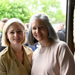 Zwei Frauen, eine mit blonden Haaren und die andere mit grauen Haaren, stehen lächelnd vor einem verschwommenen Hintergrund mit Bäumen und Menschen.