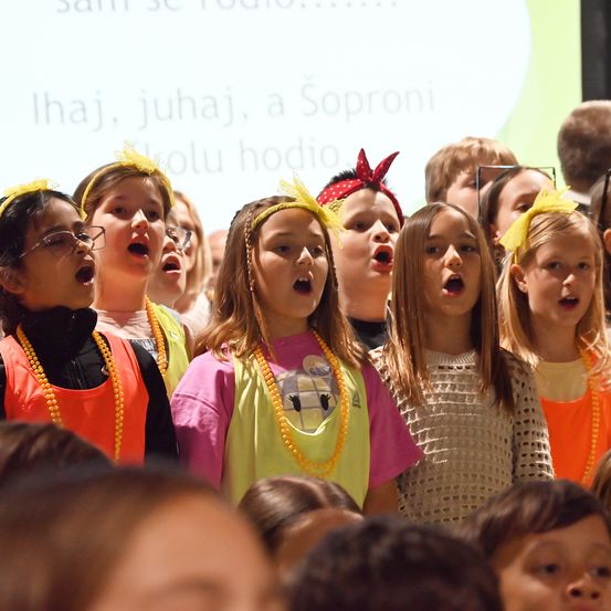 Eine Gruppe junger Mädchen mit gelben Stirnbändern und Halsketten singt gemeinsam in einem Chor, wobei eine Projektionsleinwand im Hintergrund fremdsprachigen Text anzeigt.