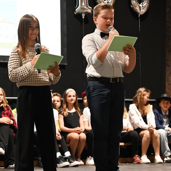 Ein Junge und ein Mädchen stehen auf der Bühne, halten grüne Papiere und Mikrofone in den Händen. Sie stehen vor einer Projektionsleinwand. Mehrere Kinder sitzen auf Bänken hinter ihnen.