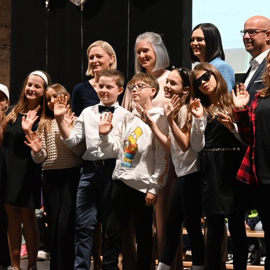 Eine Gruppe von Menschen, darunter Kinder und Erwachsene, steht gemeinsam auf einer Bühne, lächelt und winkt. Die Kinder tragen Freizeitkleidung, einige mit Brillen und Accessoires. Dahinter zeigt eine Leinwand eine Präsentation.