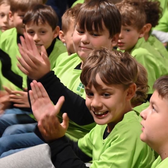 Mehrere junge Jungen in grünen Shirts sitzen zusammen, einige klatschen begeistert. Der Junge im Vordergrund lächelt und schaut etwas nach rechts.
