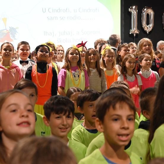 Eine Gruppe von Kindern in verschiedenen Kostümen versammelt sich für eine Aufführung, wobei eine Projektionsleinwand eine Botschaft anzeigt und Luftballons im Hintergrund zu sehen sind.