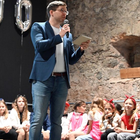 Ein Mann spricht in ein Mikrofon vor einer Gruppe von Kindern, die vor einer Felswand sitzen. Hinter ihm befindet sich ein silberner Ballon mit der Nummer 0.