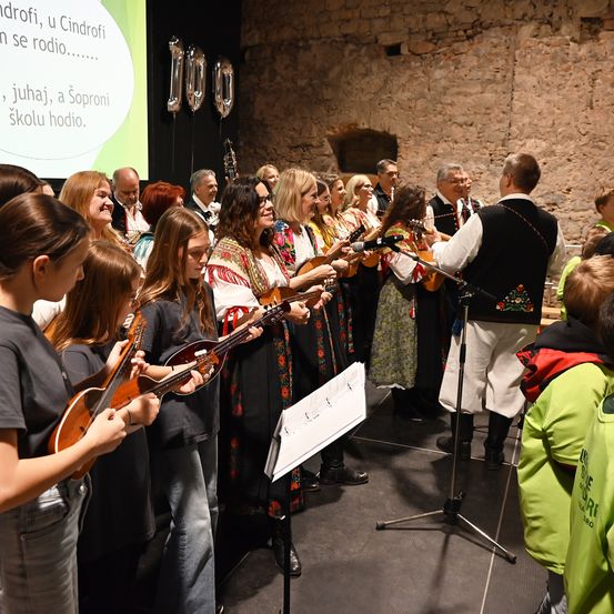 Eine Gruppe von Menschen ist versammelt, möglicherweise für eine musikalische Aufführung. Sie halten Gitarren und haben ein Mikrofon davor. Sie tragen traditionelle Kleidung. Dahinter befindet sich eine Projektionsleinwand mit Text. Dahinter hängt eine Wand mit Luftballons.