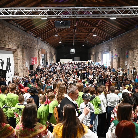 Eine große Menschenmenge versammelt sich in einem Saal, alle tragen grüne Hemden. Einige halten Gitarren, während andere Fotos machen. Ein Mann in Anzug steht in der Mitte und spricht die Menge an. Der Saal hat Steinmauern und eine Holzdecke mit Beleuchtung.