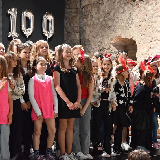 Eine Gruppe junger Mädchen, alle festlich gekleidet, steht vor einem Hintergrund mit Luftballons, die '100' anzeigen. Sie lächeln und scheinen für ein Foto zu posieren.