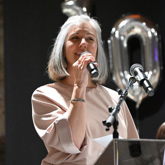 Eine Frau mit grauem Haar spricht in ein Mikrofon an einem Rednerpult und hält ein Papier in der Hand. Hinter ihr befindet sich eine große Null und ein dunkler Hintergrund.