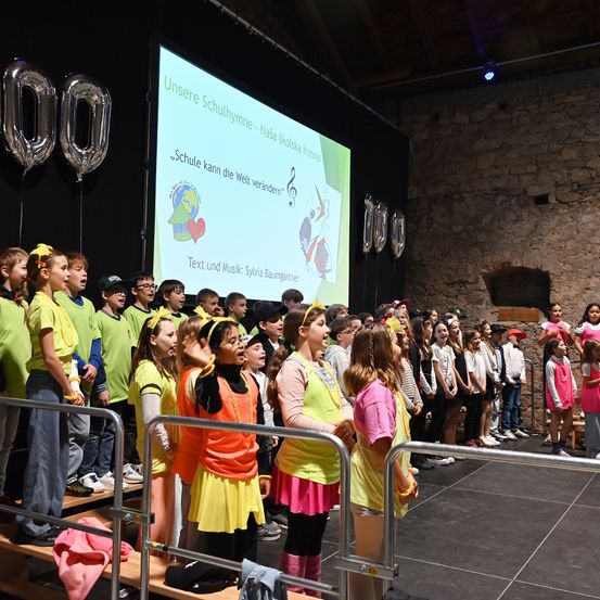 Eine Gruppe von Kindern, gekleidet in Grün, Gelb und Pink, singt vor einer Leinwand mit einem deutschen Slogan. Es gibt Luftballons und ein Hausbild auf der Leinwand.