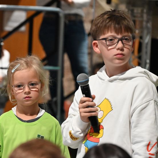 Zwei Kinder, ein Junge in einer weißen Kapuzenjacke mit Bart-Simpson-Aufdruck und ein Mädchen in einem grünen Shirt, stehen vor einem Mikrofon. Sie scheinen an einer Veranstaltung teilzunehmen, mit einer verschwommenen Person in Jeans und einem Metallgeländer im Hintergrund.