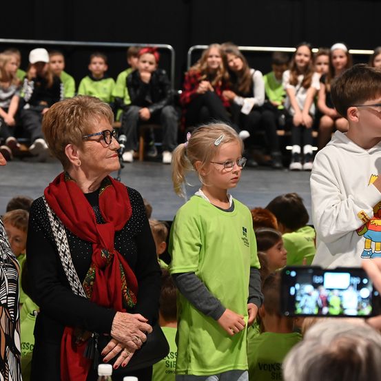 Eine ältere Frau mit Brille und einem roten Schal steht neben einem jungen Mädchen mit Zopf in Grün. Sie sehen sich eine Aufführung auf der Bühne an. Dahinter sitzen Kinder in Grün auf den Tribünen. Ein Junge in einem weißen Hoodie mit einem Simpsons-Charakter steht in der Nähe.