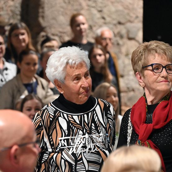 Zwei ältere Frauen, eine mit weißen Haaren und einem gemusterten Oberteil, die andere mit Brille und einem roten Schal, stehen vor einer Menschenmenge. Der Hintergrund zeigt eine Steinmauer und verschiedene Personen im Fokus.
