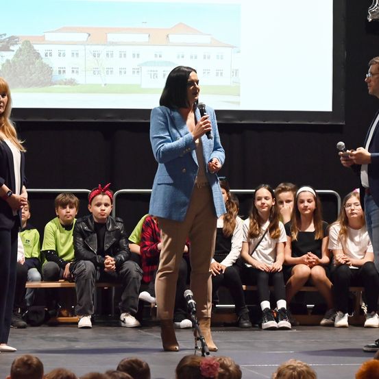 Eine Frau steht mit einem Mikrofon auf der Bühne und spricht vor einer Gruppe von Kindern und Erwachsenen. Die Kinder sitzen auf Bänken und hören aufmerksam zu. Eine große Leinwand hinter ihnen zeigt ein architektonisches Design.