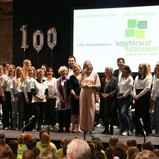 Eine Gruppe von Menschen auf der Bühne mit einer großen Leinwand im Hintergrund. Die Frau in der Mitte hält ein Mikrofon. Viele Menschen schauen von der Zuschauertribüne zu. Silberne Luftballons mit der Nummer 100 stehen hinter der Bühne.