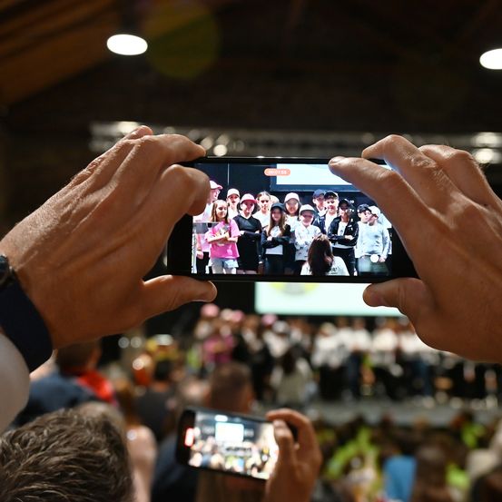 Eine Person fotografiert eine Gruppenaufnahme auf einem Smartphone, mit einer großen Menschenmenge im Hintergrund. Das Foto zeigt eine Reihe von Menschen, einige mit Mützen.
