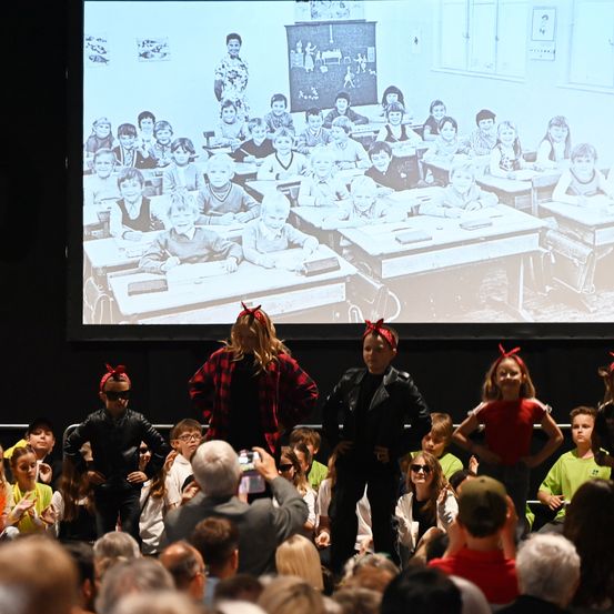 Eine Gruppe von Darstellern steht vor einem Publikum auf der Bühne, mit einem projizierten Klassenzimmer-Bild im Hintergrund. Die Darsteller sind in verschiedenen Kostümen gekleidet.