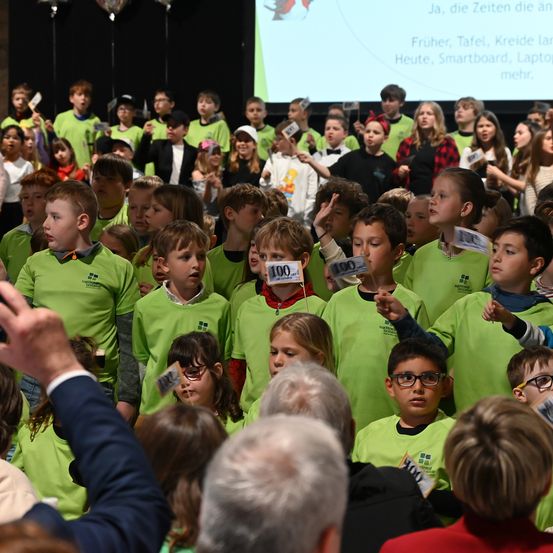 Eine Gruppe von Kindern in grünen Shirts tritt auf einer Bühne auf. Einige halten Papiere mit Zahlen. Eine große Leinwand im Hintergrund zeigt einen Vergleich von vergangenen und heutigen Werkzeugen.