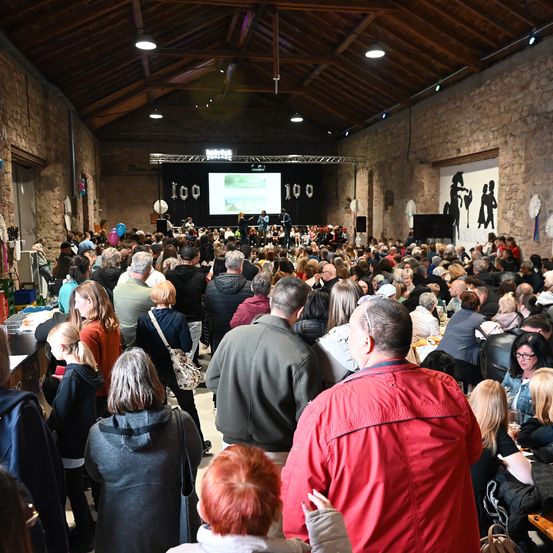 Eine große Menschenmenge ist in einem Saal versammelt und beobachtet eine Präsentation auf einer großen Leinwand. Das Publikum ist vielfältig und besteht aus Männern und Frauen. Einige sitzen, andere stehen. Es gibt Luftballons und Dekorationen im Raum.